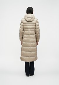 Cappotto invernale imbottito beige con cappuccio, caratterizzato da una texture lucida, trapuntatura orizzontale e una silhouette lunga che arriva sotto le ginocchia.