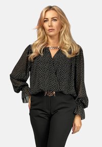 Schwarze, durchsichtige Bluse mit einem weißen, dreieckigen Muster, langen Ärmeln und einem V-Ausschnitt. Kombiniert mit schwarzen Hosen und einem metallischen Logo-Gürtel.