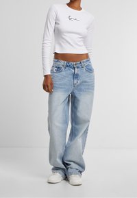 Bílé dlouhé rukávy crop top s černým logem, v kombinaci s volnými džínami se světle modrou praní a bílými teniskami, s uvolněným střihem a denimovou texturou.