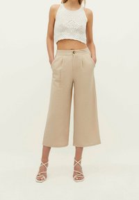 Pantalons larges beige en tissu texturé, avec deux poches avant et une fermeture à bouton, associés à un crop top blanc et des sandales à lanières.