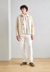 Beige canvas takki mustilla nappuloilla ja etutaskuilla, valkoisen paidan ja valkoisten housujen päällä, ruskeiden kenkien kera puulattialla.