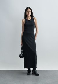 Γυναίκα με μαύρο αμάνικο μάξι φόρεμα με ριγέ υφή, κρατώντας μια μικρή μαύρη τσάντα και φοράει μαύρα μποτάκια μέχρι τον αστράγαλο, στέκεται μπροστά σε έναν μονόχρωμο γκρι τοίχο.