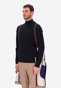 Maglione a collo alto navy, misto lana, dal design aderente. Cappotto beige e blu appoggiato sul braccio, con piping a contrasto e dettagli sulle tasche.