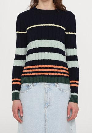 Personne portant un pull torsadé bleu marine avec des rayures horizontales jaunes, bleu clair, orange et vert, associé à une jupe en jean bleu clair.