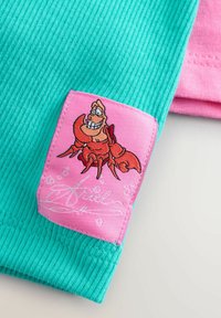 Turkist ribbet stof med et lyserødt patch, der har en broderet tegneseriekrabbe. Patchen inkluderer bobler og signaturdetalje.
