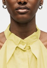 Κοντινό πλάνο ενός ατόμου που φοράει ασημένια κρίκους σκουλαρίκια και ανοιχτό κίτρινο τοπ με λαιμόκοψη halter και κολάρο με κουμπιά.