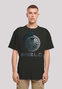 Svart bomullst-skjorte med grafisk design som har et sirkulært emblemet og teksten "S.H.I.E.L.D." i metallisk stil, korte ermer, avslappet passform.
