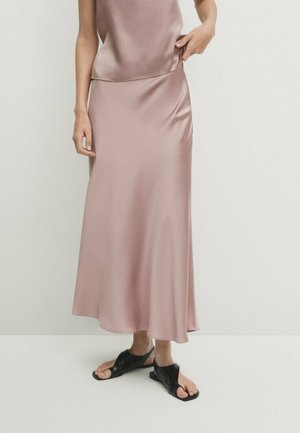 Kobieta w jasnoróżowej satynowej spódnicy midi i pasującym topie, zestawiona z czarnymi płaskimi sandałami z odkrytymi palcami, stojąca na jednolitym tle.