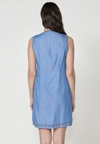Robe en denim sans manches de couleur bleu clair, avec une coupe droite, une finition texturée, et un délicat broderie noire en zigzag le long de l'ourlet.