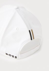 White baseball cap with a curved brim, adjustable strap at the back, black BOSS logo, and three accent stripes in black, white, and gold.