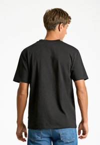 Maglietta nera a maniche corte con scollo rotondo, tessuto a trama, e vestibilità morbida, indossata con jeans blu, vista da dietro.