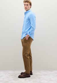 Camicia azzurro chiaro con bottoni e taschino sul petto, abbinata a pantaloni marroni e scarpe in pelle marrone. Il modello posa su uno sfondo neutro.