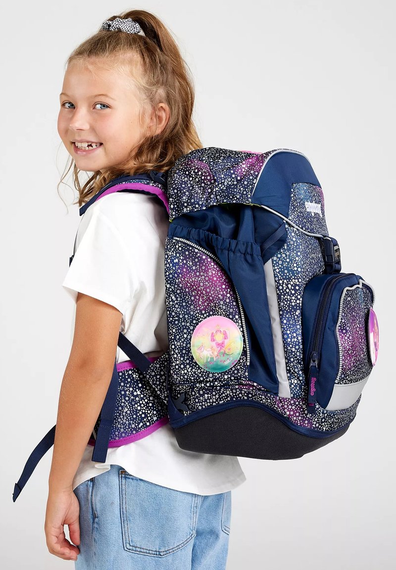 Lächendes junges Mädchen mit hellem Haar, das einen Rucksack mit Galaxie-Muster, reflektierenden Patches und rosa Besatz über einem weißen Shirt und Jeans trägt.