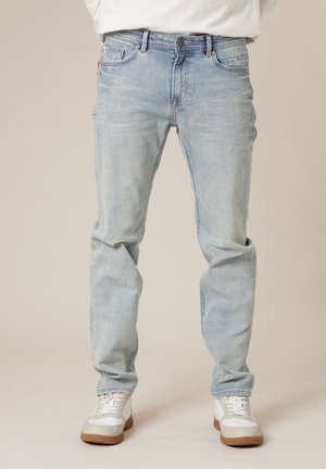 Mann trägt hellblaue Jeans und weiße Turnschuhe und steht vor einem einfarbigen Hintergrund.