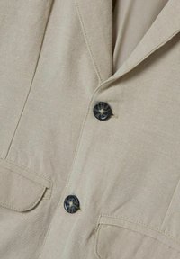 Blazer beige con colletto a punta, due tasche applicate e dettagli in bottoni scuri. Il materiale presenta una texture fine e una lavorazione sottile.