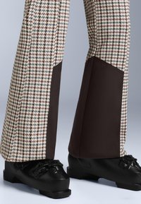 Nohavice so vzorom houndstooth v béžovej, hnedej a čiernej farbe s kontrastnými tmavými textilnými akcentmi; v kombinácii s čiernymi lyžiarskymi topánkami s prackami.