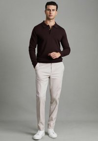 Maglione bordeaux scuro con colletto e tre bottoni, abbinato a pantaloni beige chiaro e sneakers bianche su uno sfondo neutro.