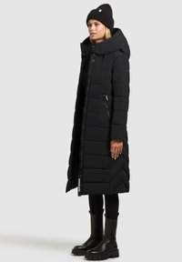 Vrouw in een lange zwarte gewatteerde jas met capuchon, zwarte beanie, zwarte leggings en dikke zwarte laarzen, staande tegen een grijze achtergrond.