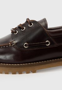 Chaussure en cuir brun foncé avec une forme arrondie, œillets en métal argenté et lacets marron. Présente une semelle en caoutchouc texturée avec un profil de crampons prononcé.