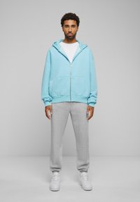  világoskék cipzáros kapucnis felső, elülső zsebbel, szürke melegítőnadrággal és fehér sportcipővel párosítva. Egyszerű, laza sportos öltözet.