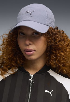 Kobieta z kręconymi włosami, ubrana w jasnoszary daszek Puma oraz czarną kurtkę Puma z białymi akcentami, zwrócona przodem na tle szarego tła.