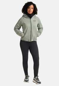 Giacca a zip verde chiaro con cappuccio, base nera e pantaloni neri. Le caratteristiche includono polsini elastici e tasche. Calzature multicolori.
