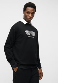 Άνδρας με μαύρο φούτερ, λευκά αφαιρετικά γυαλιά ηλίου και κείμενο «KARL LAGERFELD», συνδυασμένος με μαύρο παντελόνι και πουκάμισο με λευκό γιακά.