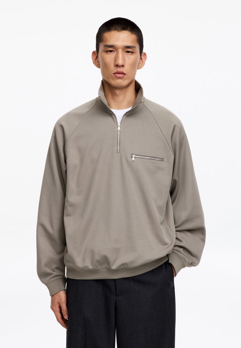 Homme portant un pull taupe à fermeture éclair quart, avec une poche zippée poitrine et un pantalon foncé, debout avec une main dans la poche.