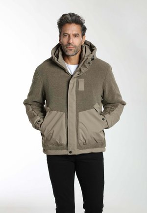 Homme debout les mains dans les poches portant une veste à capuche en polaire marron et nylon avec un pantalon noir sur un fond uni.