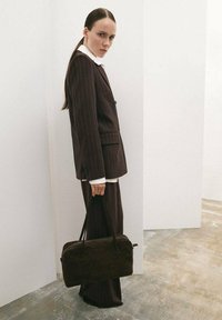 Traje marrón a rayas con chaqueta entallada y pantalones de pierna ancha. Camisa blanca con cuello debajo. Sosteniendo un bolso negro texturizado. Fondo neutro.