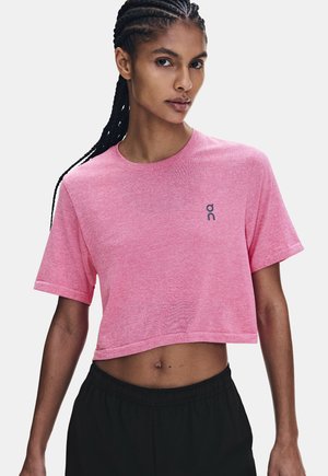 Frau mit geflochtenem Haar, die ein pinkfarbenes, kurzes T-Shirt mit kleinem Logo und schwarze Hosen trägt, steht vor einem schlichten hellen Hintergrund.