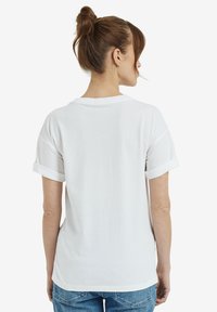 Frau mit braunen Haaren im Dutt, die ein schlichtes weißes T-Shirt mit kurzen Ärmeln und blaue Jeans trägt, von hinten gegen einen weißen Hintergrund abgebildet.