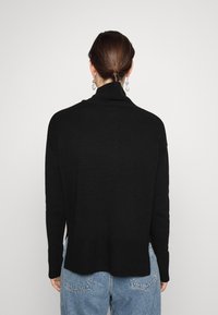 Black knitted sweater with a high collar, ribbed cuffs, and a slightly extended back hem, paired with light blue denim jeans.