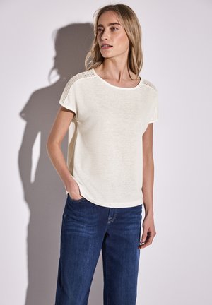 Crème korte mouw top met een relaxte pasvorm, voorzien van gestructureerde breiaccenten op de schouders. Gecombineerd met donkerblauwe jeans.