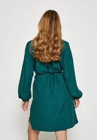 Mornarsko zelena obleka z teksturirano površino, dolgimi nabranimi rokavi in zoženim pasom. Ima stranske razpoke in sproščen kroj.