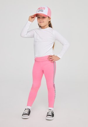 Leggings rosa con strisce laterali, abbinati a una maglietta a maniche lunghe bianca e un cappellino rosa e bianco con il testo "PARIS". Sneakers nere.
