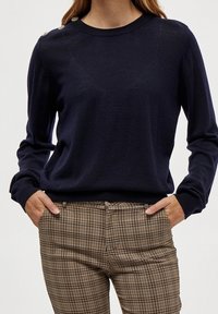 Marinblå stickad tröja med långa ärmar och axelknappdetaljer, tillsammans med rutig byxa i bruna och beige nyanser.