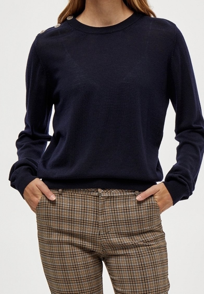 Marinblå stickad tröja med långa ärmar och axelknappdetaljer, tillsammans med rutig byxa i bruna och beige nyanser.