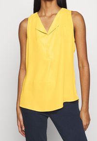 Donna che indossa un top giallo senza maniche con colletto a V, abbinato a pantaloni scuri blu navy, su uno sfondo chiaro uniforme.