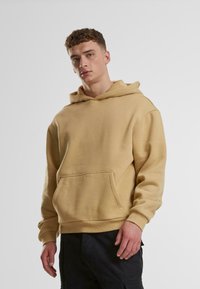 Felpa in cotone beige con tasca frontale, cappuccio con cordino e polsini a coste. Il modello la indossa con pantaloni cargo scuri.