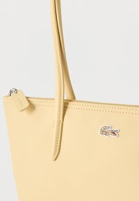 Sac fourre-tout jaune pâle texturé avec des bretelles fines, fermeture éclair et petit logo de crocodile brodé à l'avant.