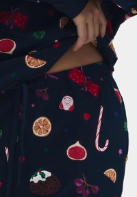 Conjunto de pijama azul marino con patrones coloridos de frutas y motivos navideños, incluyendo naranjas, bastones de caramelo y regalos. Material suave, corte relajado.