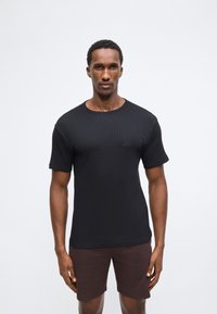 Homme debout portant un T-shirt noir à manches courtes et un short marron foncé devant un fond clair uni.