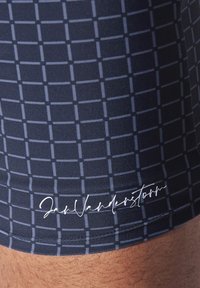 Navy swim shorts featuring a grid pattern in lighter blue, made of smooth fabric with an embroidered white logo along the hem.