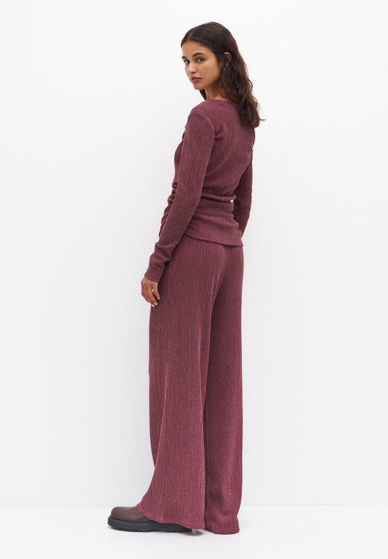 Top a maniche lunghe a coste color bordeaux abbinato a pantaloni a gamba larga; texture liscia e design aderente; indossato con scarpe scure a suola bassa marroni.