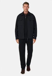 Črna zip-up jakna z dvema sprednjima žepoma, v kombinaciji s črno srajco z dolgimi rokavi in ujemajočimi hlačami. Model nosi rjave superge.