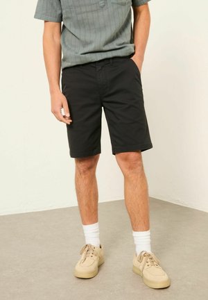 Mann trägt schwarze knielange Shorts, graues kurzärmeliges Hemd, weiße Socken und beige Turnschuhe und steht auf einem grauen Boden.