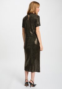 Femme aux cheveux blonds portant une robe midi noire et dorée en sequins et des escarpins noirs, se tenant devant un fond blanc uni.