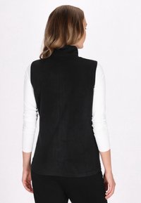 Gilet nero senza maniche realizzato in tessuto morbido e strutturato, con colletto alto, abbinato a una camicia bianca a maniche lunghe sotto.