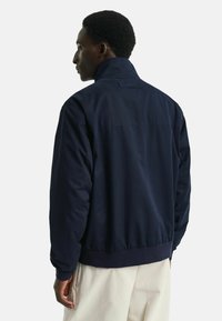 Giacca blu navy con colletto alto, caratterizzata da orli e polsini a coste. Tessuto liscio, design semplice, con un logo minimale sul retro.